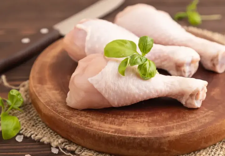 Trois cuisses de poulet cru sur une planche en bois, garnies de basilic. Un couteau est visible en arrière-plan. Mon chien adore le poulet cru.