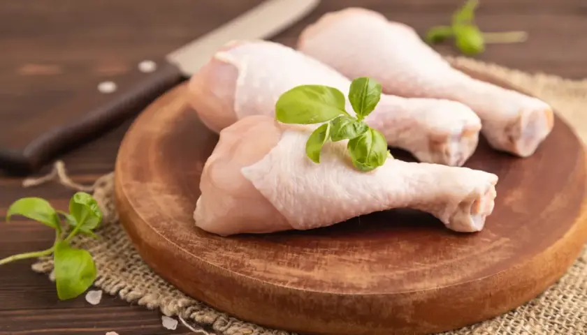 Trois cuisses de poulet cru sur une planche en bois, garnies de basilic. Un couteau est visible en arrière-plan. Mon chien adore le poulet cru.