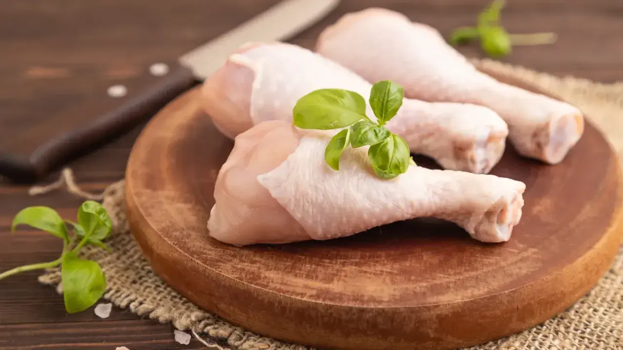 Trois cuisses de poulet cru sur une planche en bois, garnies de basilic. Un couteau est visible en arrière-plan. Mon chien adore le poulet cru.
